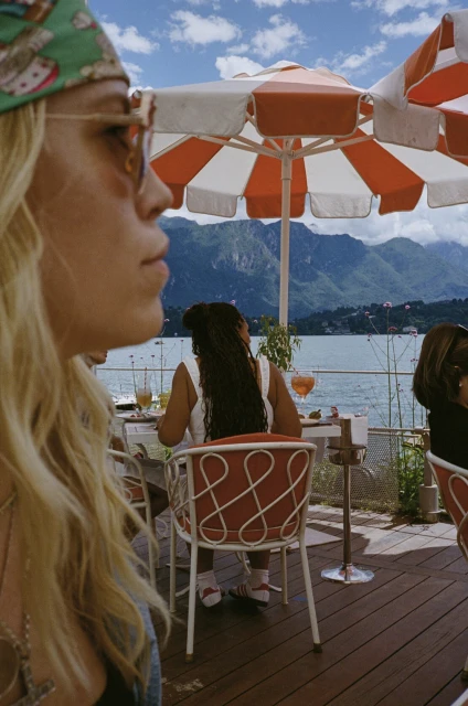 Outdoor cafe scene featuring two women enjoying beverages beneath a striped umbrella with a mountain and lake backdrop, summer vibe.