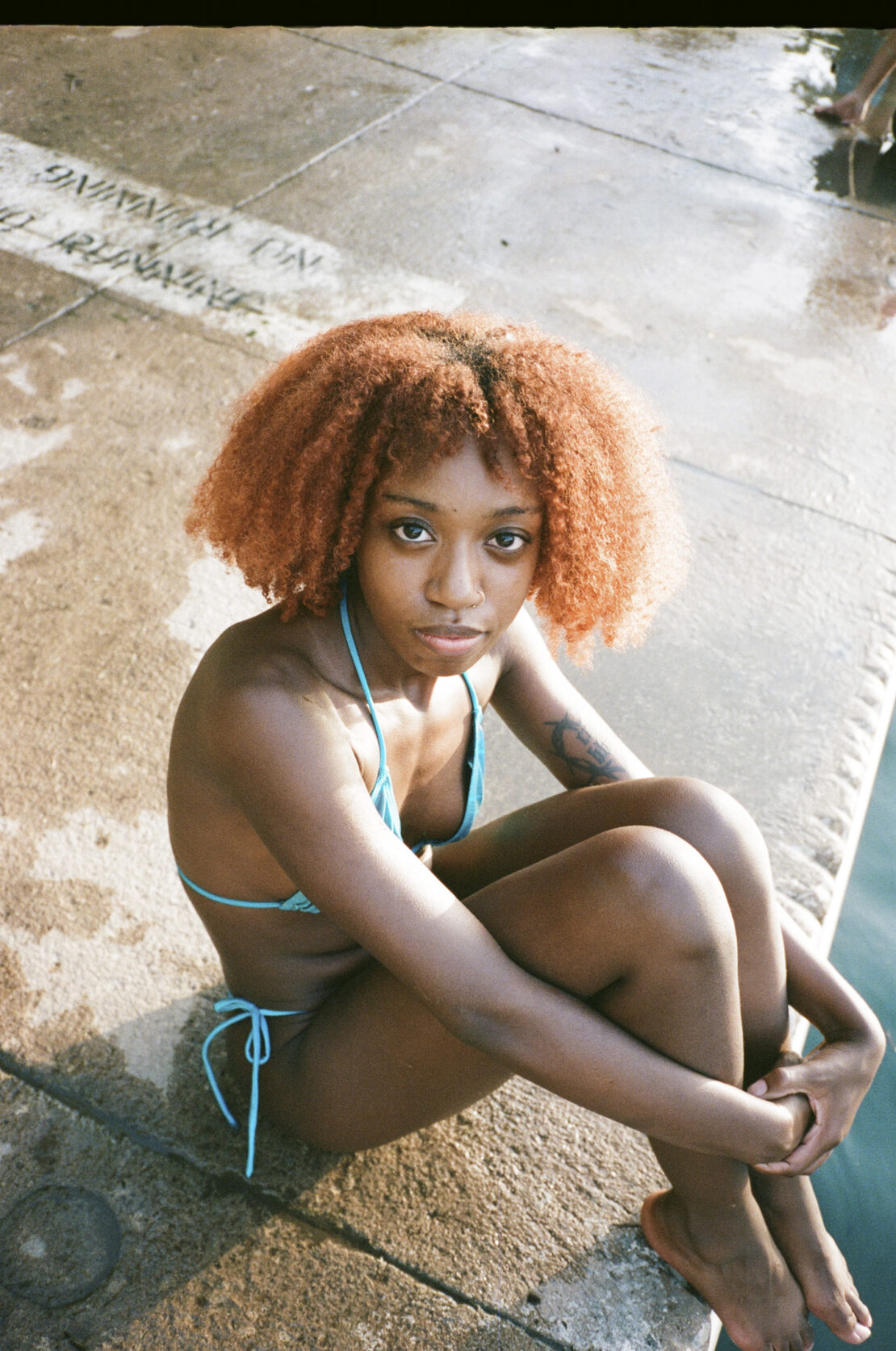 Portrait of a young Black woman with afro, posing in a teal bikini on a sunny day, seated near concrete with water nearby.
