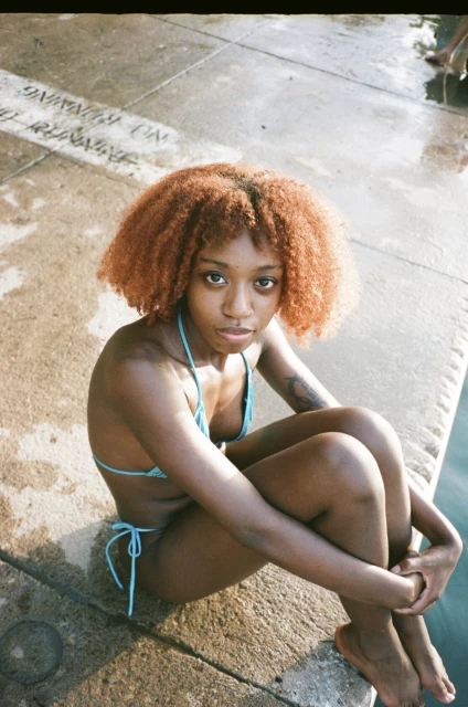 Portrait of a young Black woman with afro, posing in a teal bikini on a sunny day, seated near concrete with water nearby.