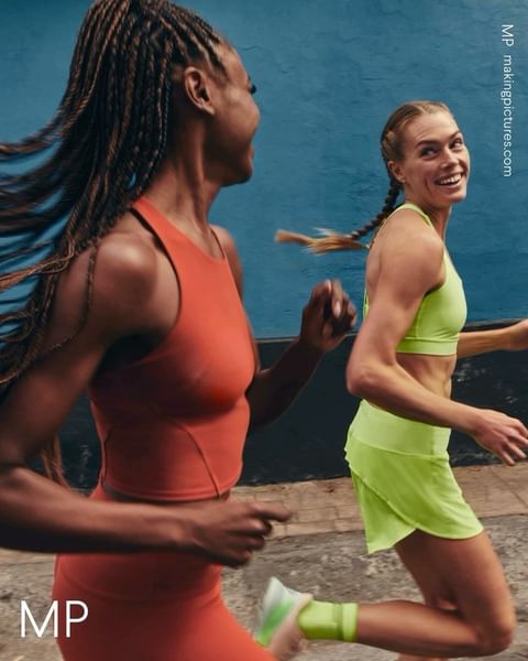 Two women in athletic wear are running outdoors on a sunny day, smiling. One wears orange, the other lime green activewear during a run.