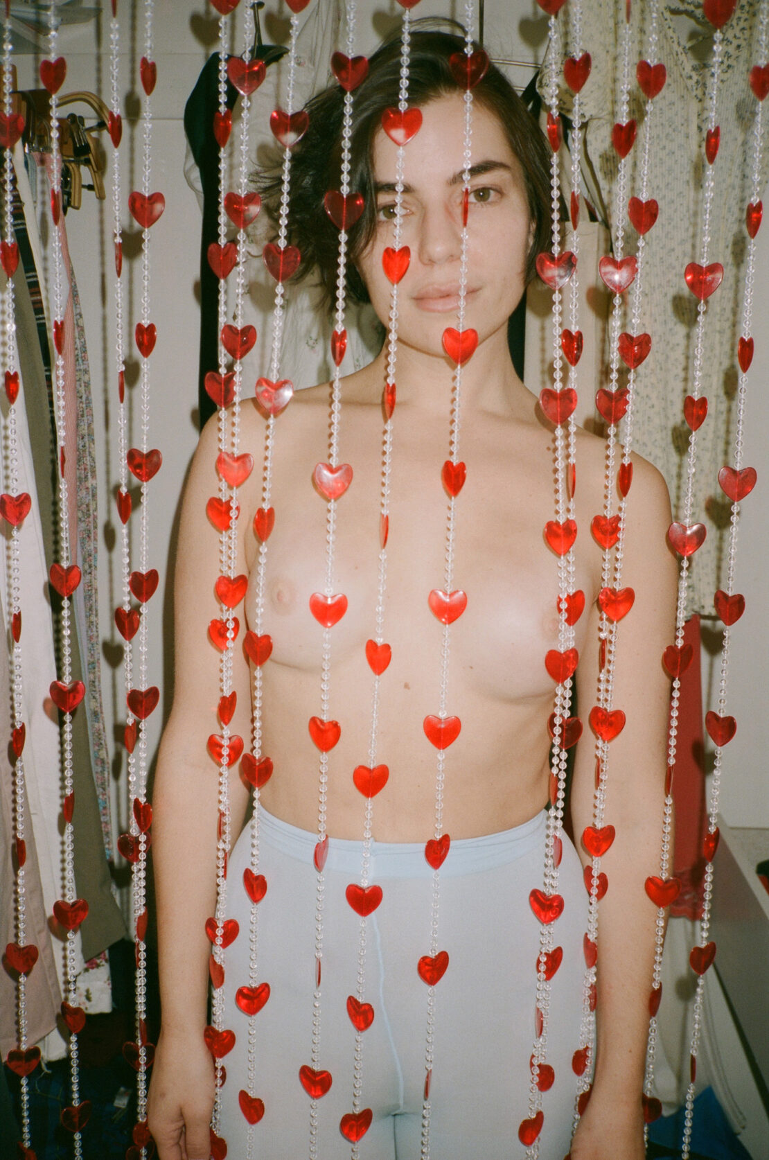 Adult female posing behind a beaded curtain of red hearts. Light blue bottoms. Indoor photo studio setting. Focus on the subject.