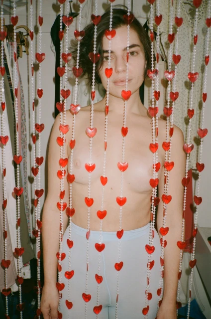 Adult female posing behind a beaded curtain of red hearts. Light blue bottoms. Indoor photo studio setting. Focus on the subject.