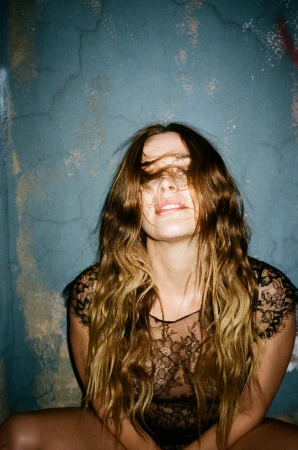 Woman with long wavy brown hair covering her face wearing a black lace top against a teal cracked wall