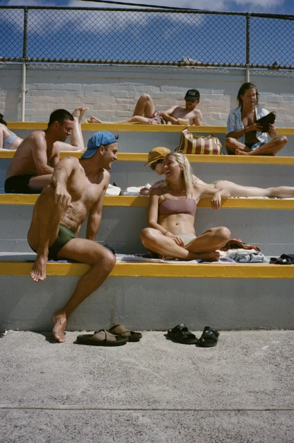 People relaxing on concrete bleachers on a sunny day. A shirtless man with a blue cap sits next to a woman in a bikini top and bottoms.