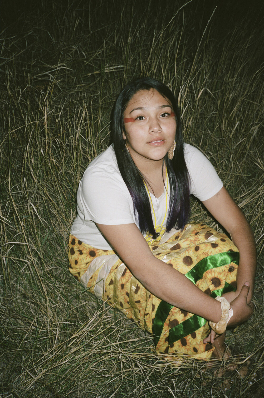 Young Indigenous woman with traditional makeup and sunflower skirt poses in a grassy field, looking directly at the camera.