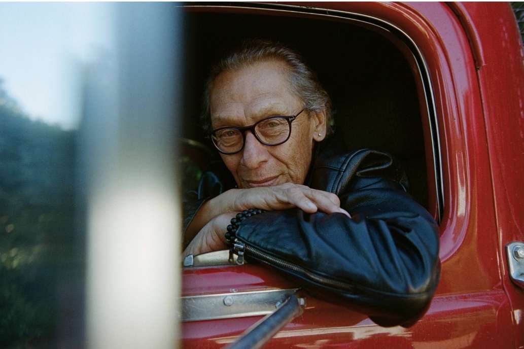 Older adult with glasses and black leather jacket leans out of the window of a red pickup truck looking at the camera.