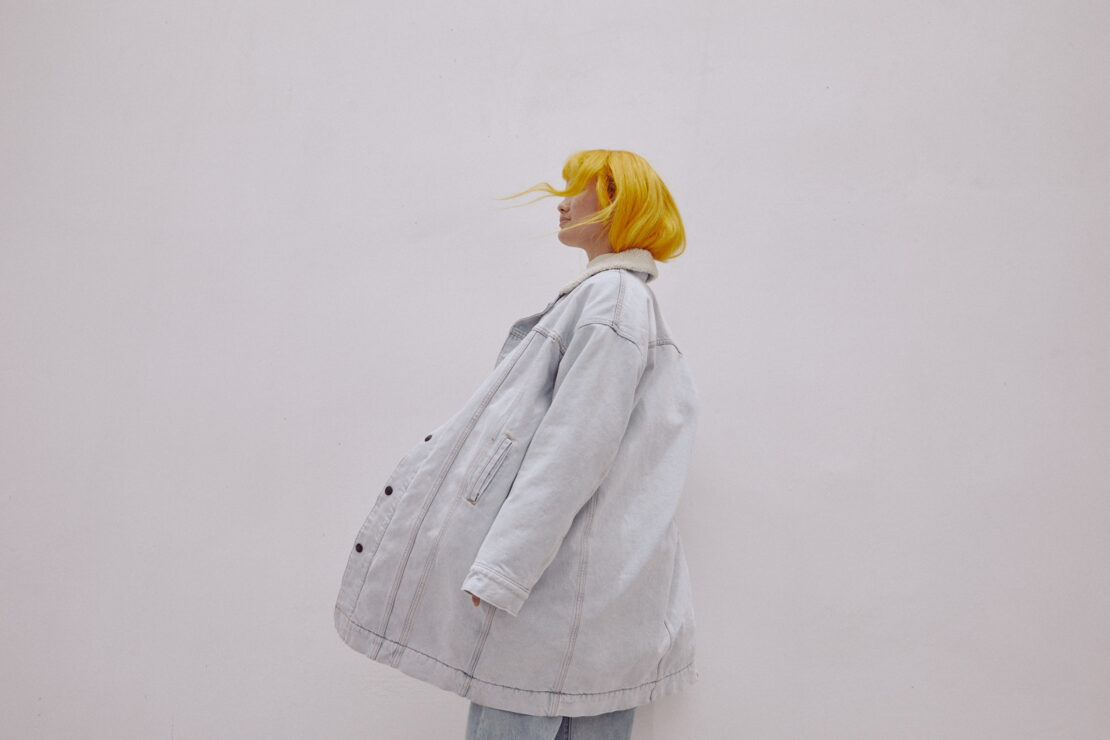 A woman with yellow hair stands in a light-blue denim jacket against a plain white background in a fashion portrait, looking off to the side.