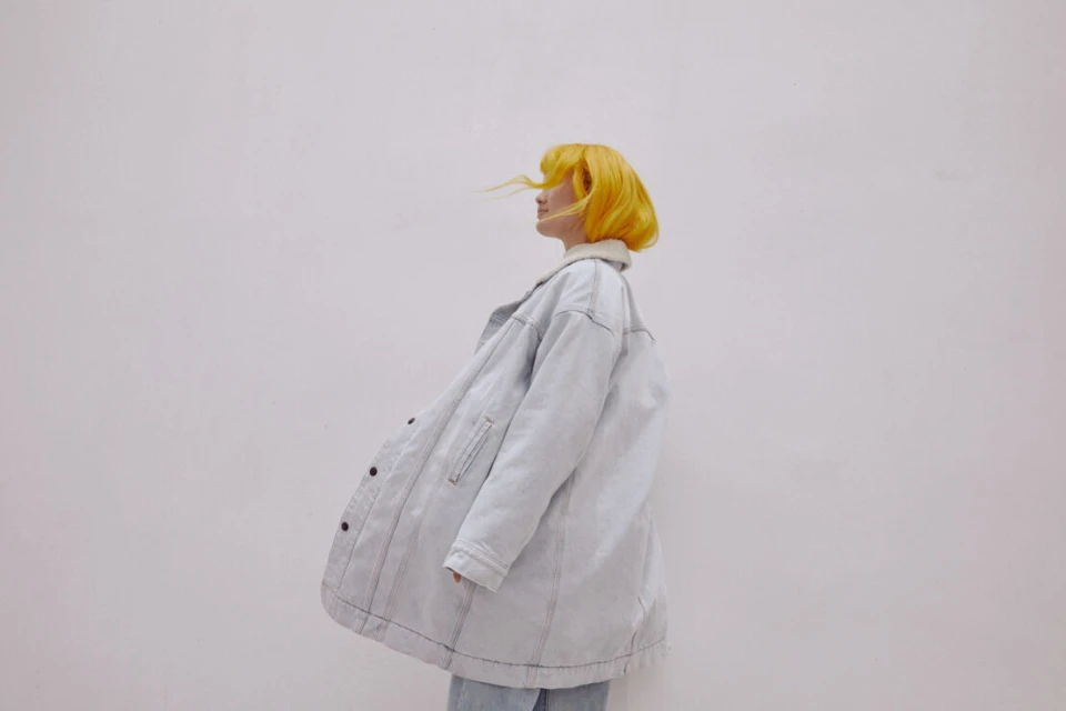 A woman with yellow hair stands in a light-blue denim jacket against a plain white background in a fashion portrait, looking off to the side.