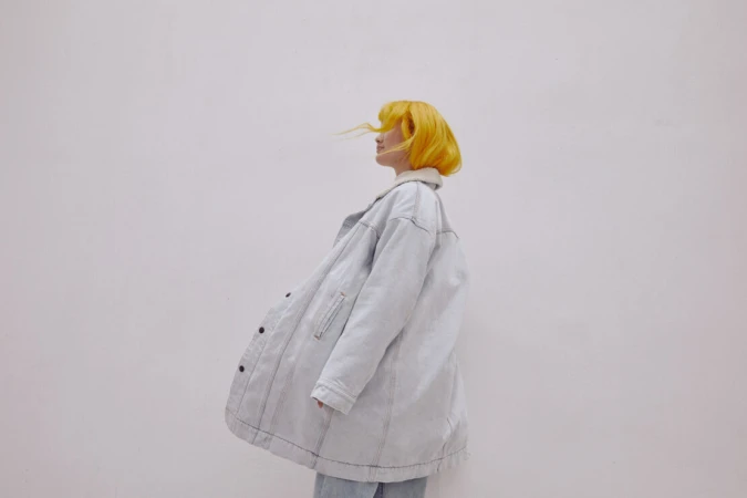 A woman with yellow hair stands in a light-blue denim jacket against a plain white background in a fashion portrait, looking off to the side.
