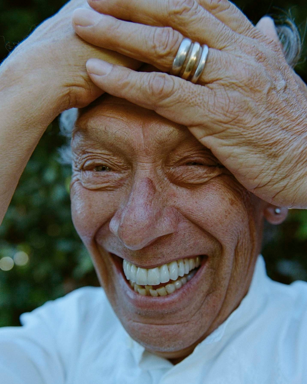 Close-up portrait of a smiling elderly adult woman with her hands near her face, wearing a white shirt, outdoors with green foliage.
