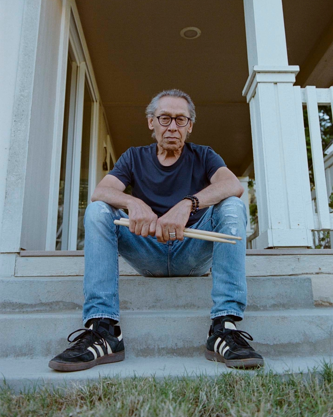 Elderly man with glasses and drumsticks sits on steps of a house. He wears a t-shirt, jeans, and black sneakers with white stripes.