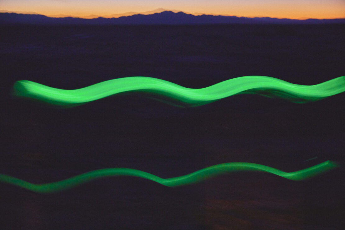Abstract image with two wavy, horizontally-oriented green light trails against a blurred background of mountains at dusk.