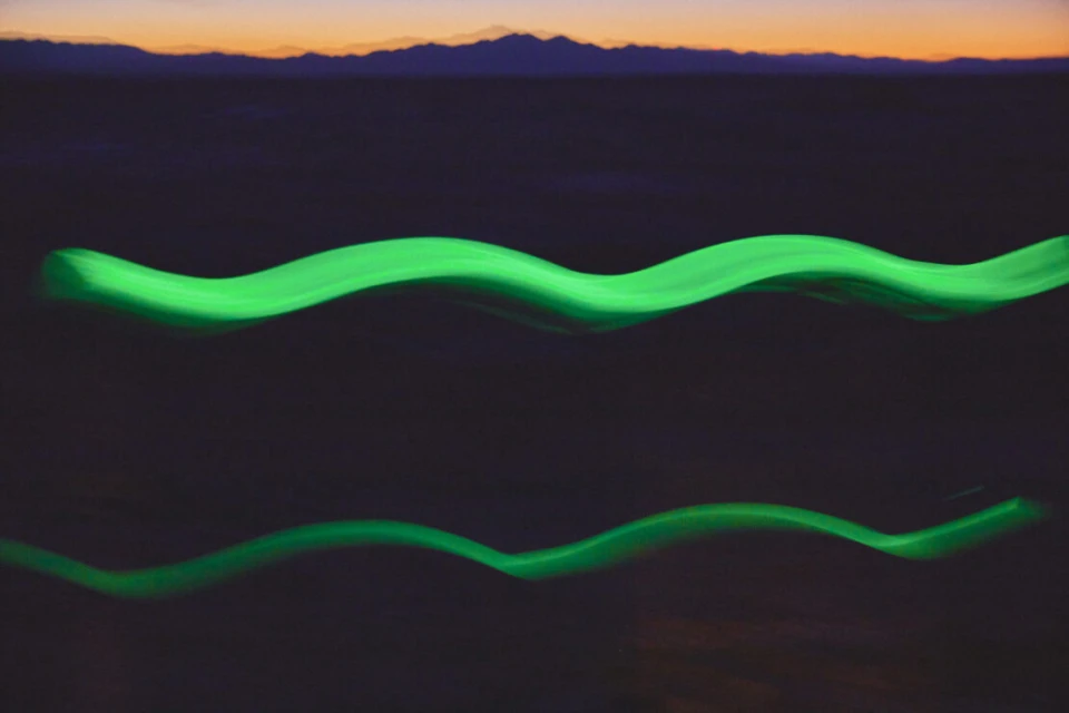 Abstract image with two wavy, horizontally-oriented green light trails against a blurred background of mountains at dusk.