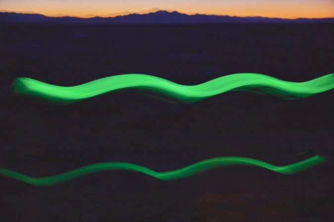 Abstract image with two wavy, horizontally-oriented green light trails against a blurred background of mountains at dusk.