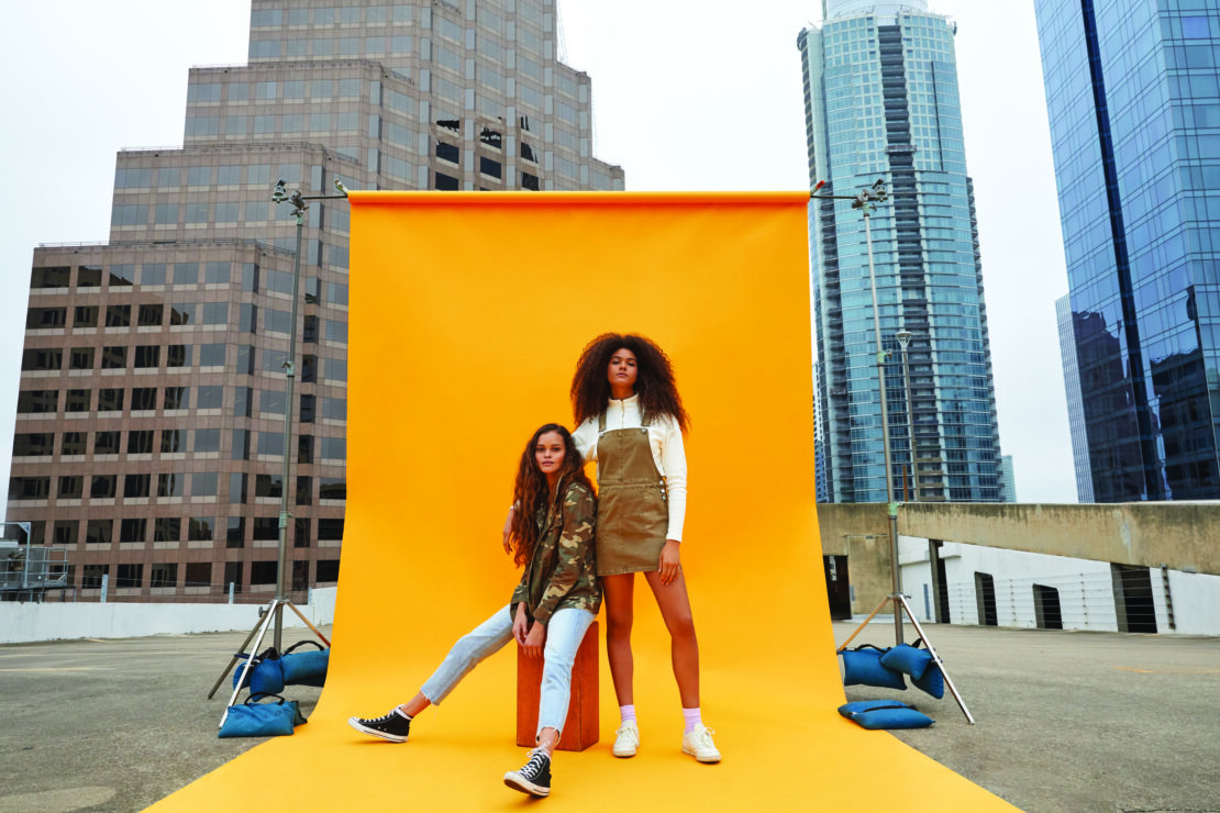 Two women pose in front of a bright yellow backdrop on a rooftop, with urban buildings creating an interesting city skyline.