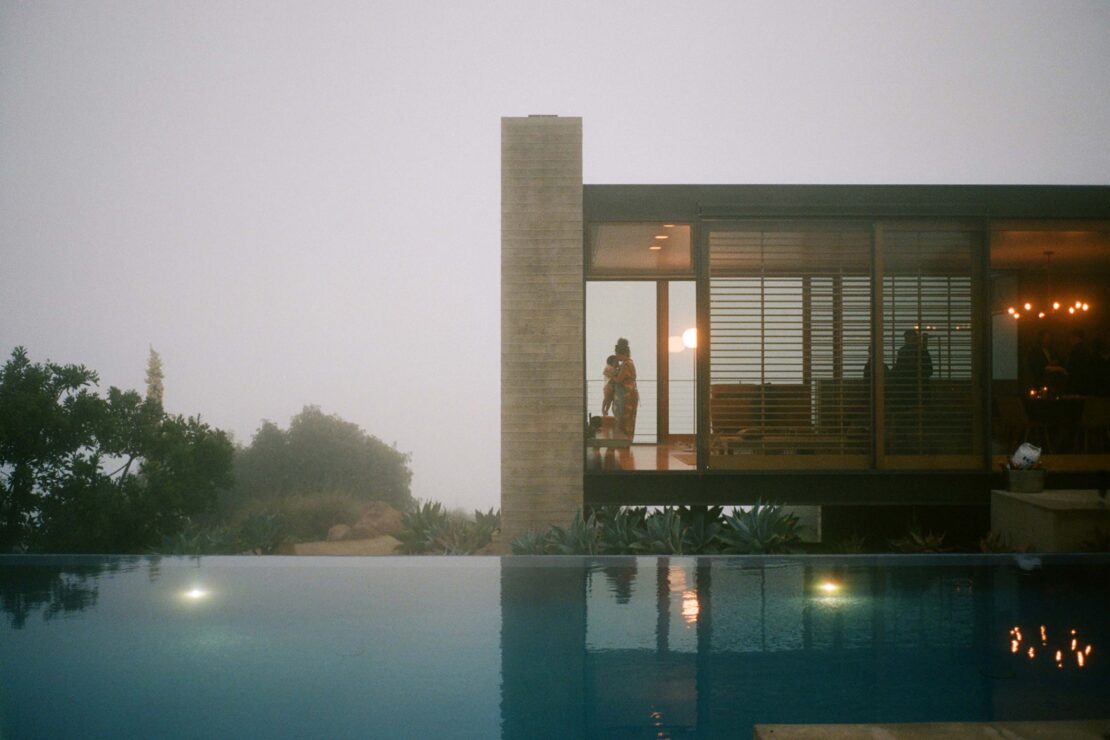 Exterior view of modern home by a pool with two people standing at sliding glass doors, reflecting the sun.