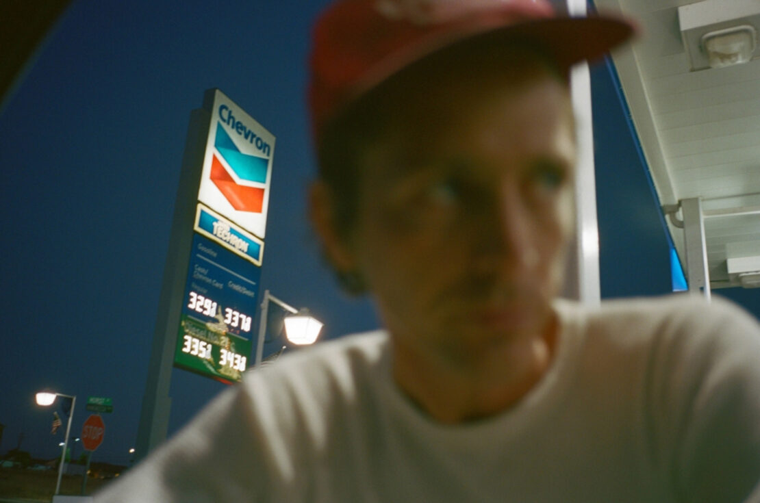 Man in a red cap stands in the foreground with a Chevron gas station sign and illuminated fuel prices visible at night.