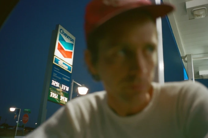 Man in a red cap stands in the foreground with a Chevron gas station sign and illuminated fuel prices visible at night.
