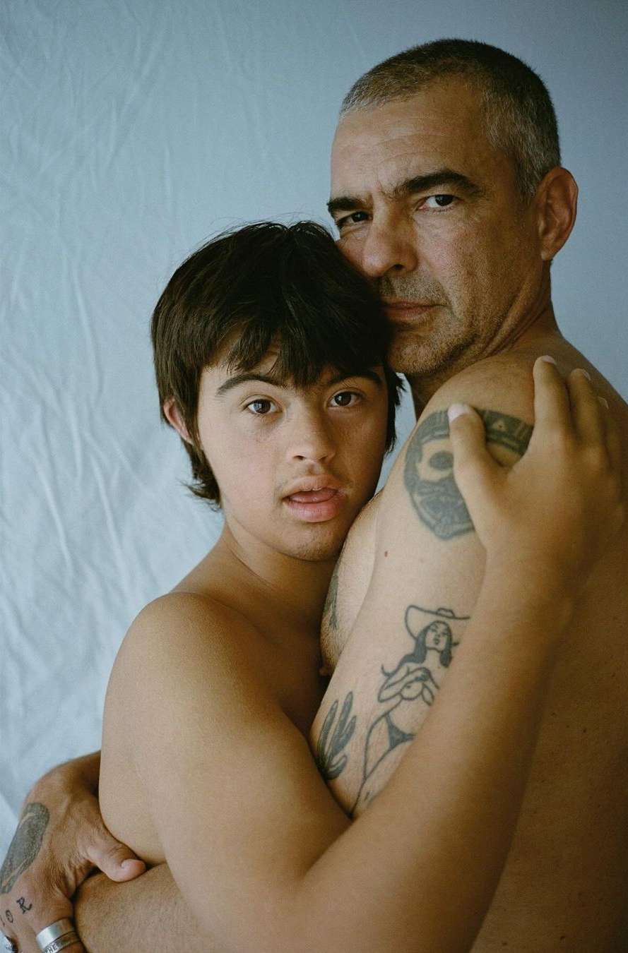 Close-up portrait of a shirtless adult male embracing a young shirtless adult male with Down syndrome against a light blue background.