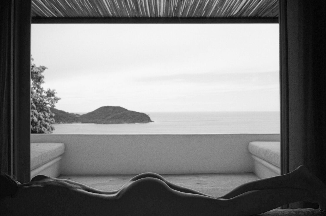 Monochrome shot of a reclining nude woman overlooking the ocean from a terrace, featuring a landscape with an island in the background.