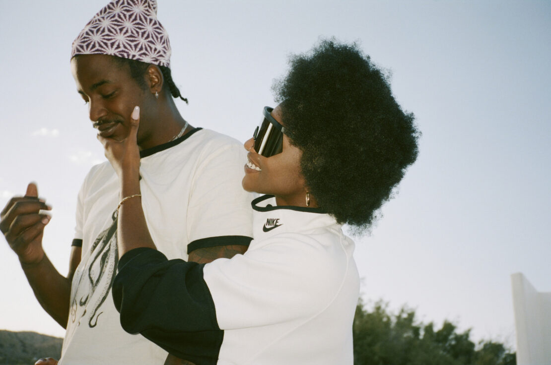 A smiling man and woman are closely posed outdoors on a sunny day. The woman, in sunglasses, touches the man's face affectionately.