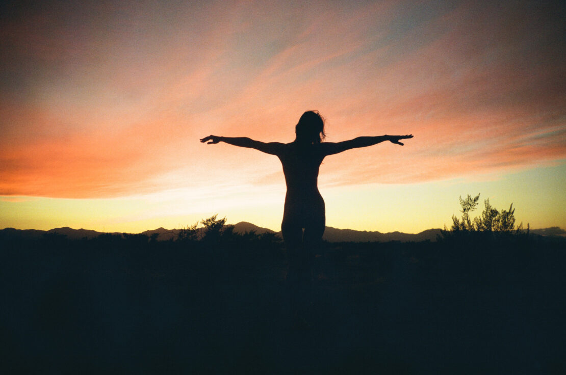 Silhouette of a slender woman with arms outstretched against a vibrant sunset sky over a dark landscape of brush and low mountains.