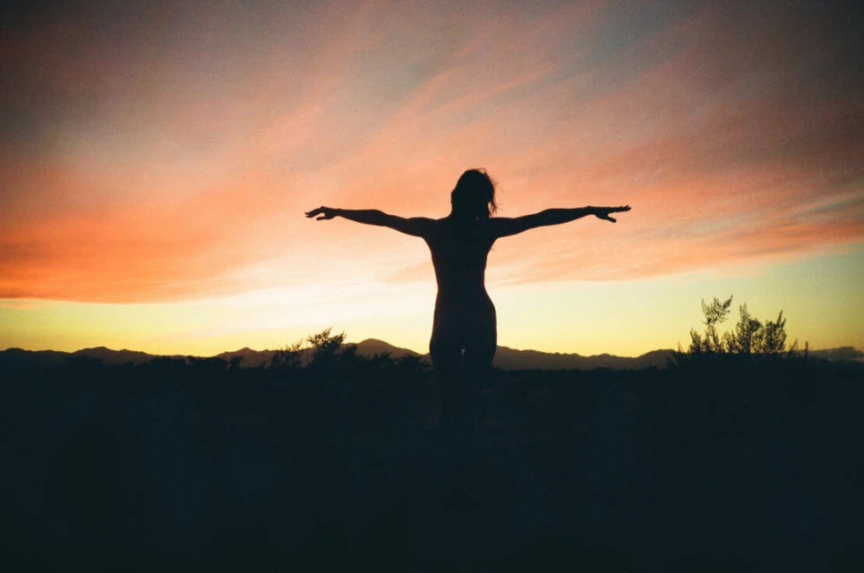 Silhouette of a slender woman with arms outstretched against a vibrant sunset sky over a dark landscape of brush and low mountains.