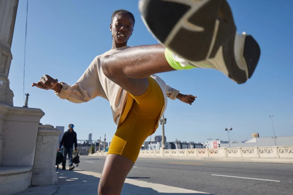 Active woman kicks leg up on a city bridge, wearing a beige top and yellow athletic shorts. Blue sky and urban buildings in background.