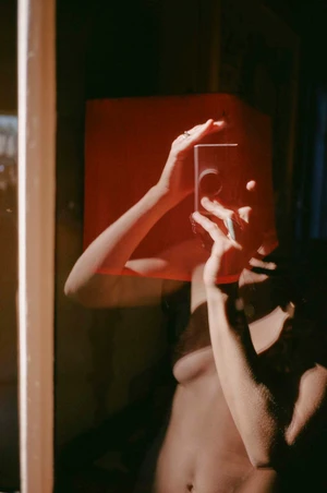 Person holding a compact camera in front of a mirror with a red lampshade reflected in the glass.