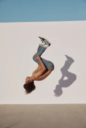 Shirtless man with curly hair performing a backflip wearing cuffed blue jeans and sneakers against a white wall.