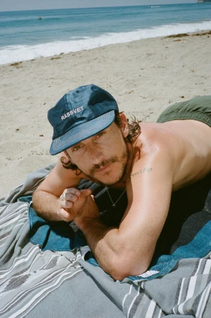 Man lying on a beach towel wearing a dark blue baseball cap with white text, positioned against an ocean and sandy beach background.