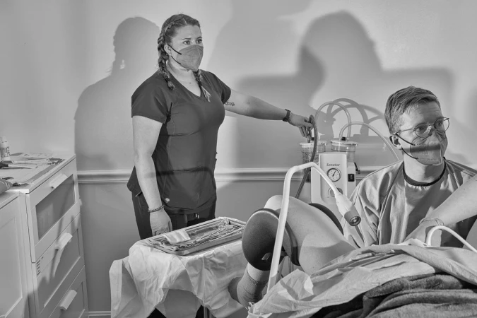 Two healthcare workers in scrubs and masks are performing a medical procedure indoors using medical equipment and tools, with a neutral background.