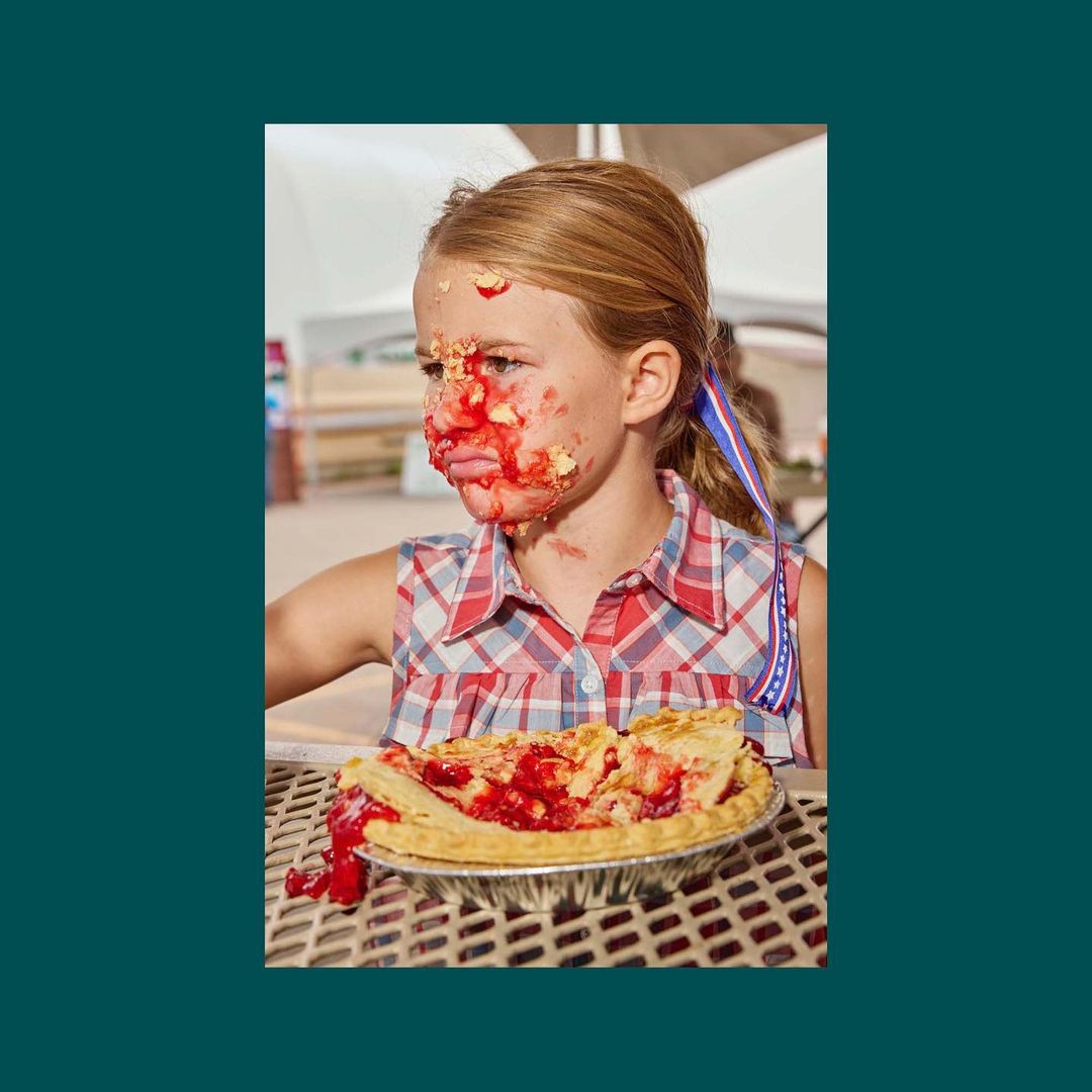 A girl with a red and white checkered shirt and a red, white, and blue ribbon in her hair has pie filling smeared on her face.
