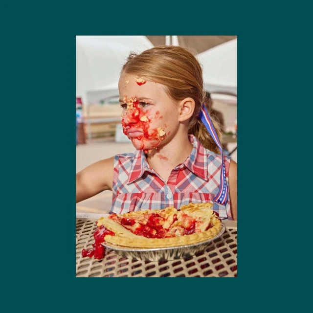 A girl with a red and white checkered shirt and a red, white, and blue ribbon in her hair has pie filling smeared on her face.