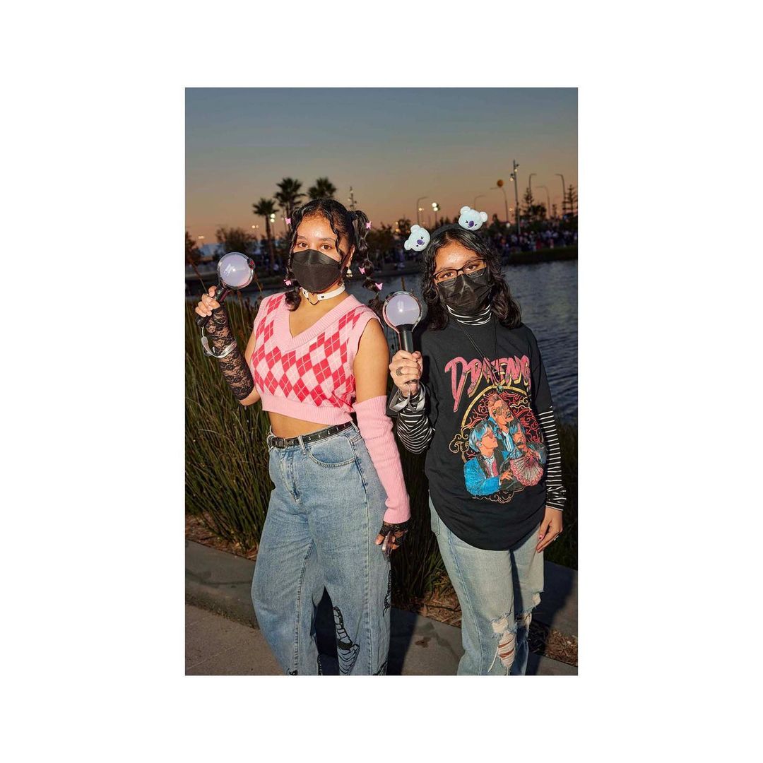 Two young women pose outdoors at dusk wearing face masks, jeans, and casual tops, each holding a light stick in front of a lake.