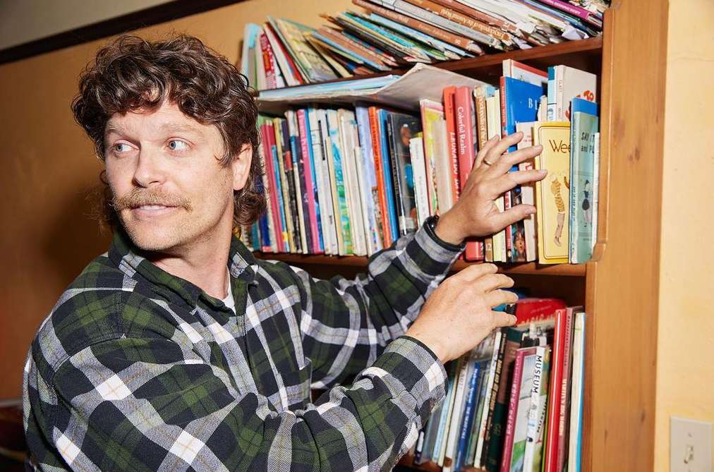Adult male with mustache and curly hair reaching into a bookshelf filled with colorful books in an indoor environment, eye level.