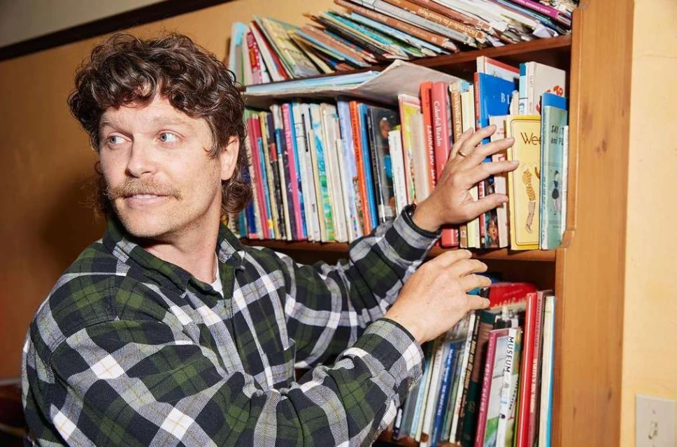 Adult male with mustache and curly hair reaching into a bookshelf filled with colorful books in an indoor environment, eye level.