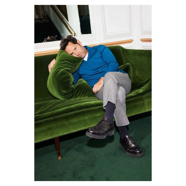 Adult male lounging on a green velvet sofa in an indoor setting, wearing a blue shirt, grey pants, and black dress shoes, looking relaxed.