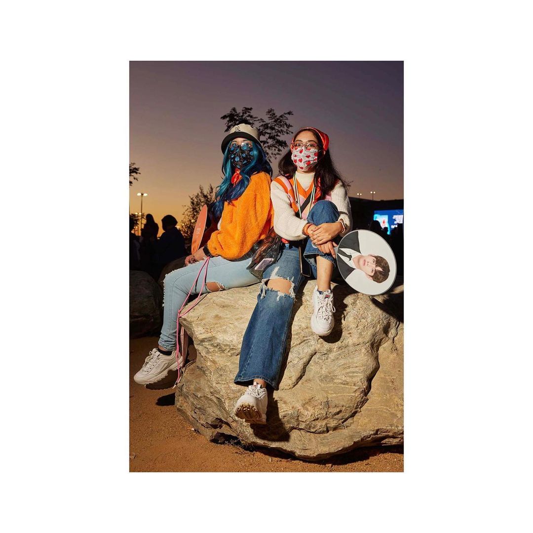 Two women in face masks sit on a rock outdoors at dusk, one holding a fan with a man's image. The women wear casual clothing and hats.