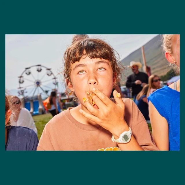 A child with brown hair and a tan t-shirt is eating from their fingers at an outdoor event with a ferris wheel in the background.