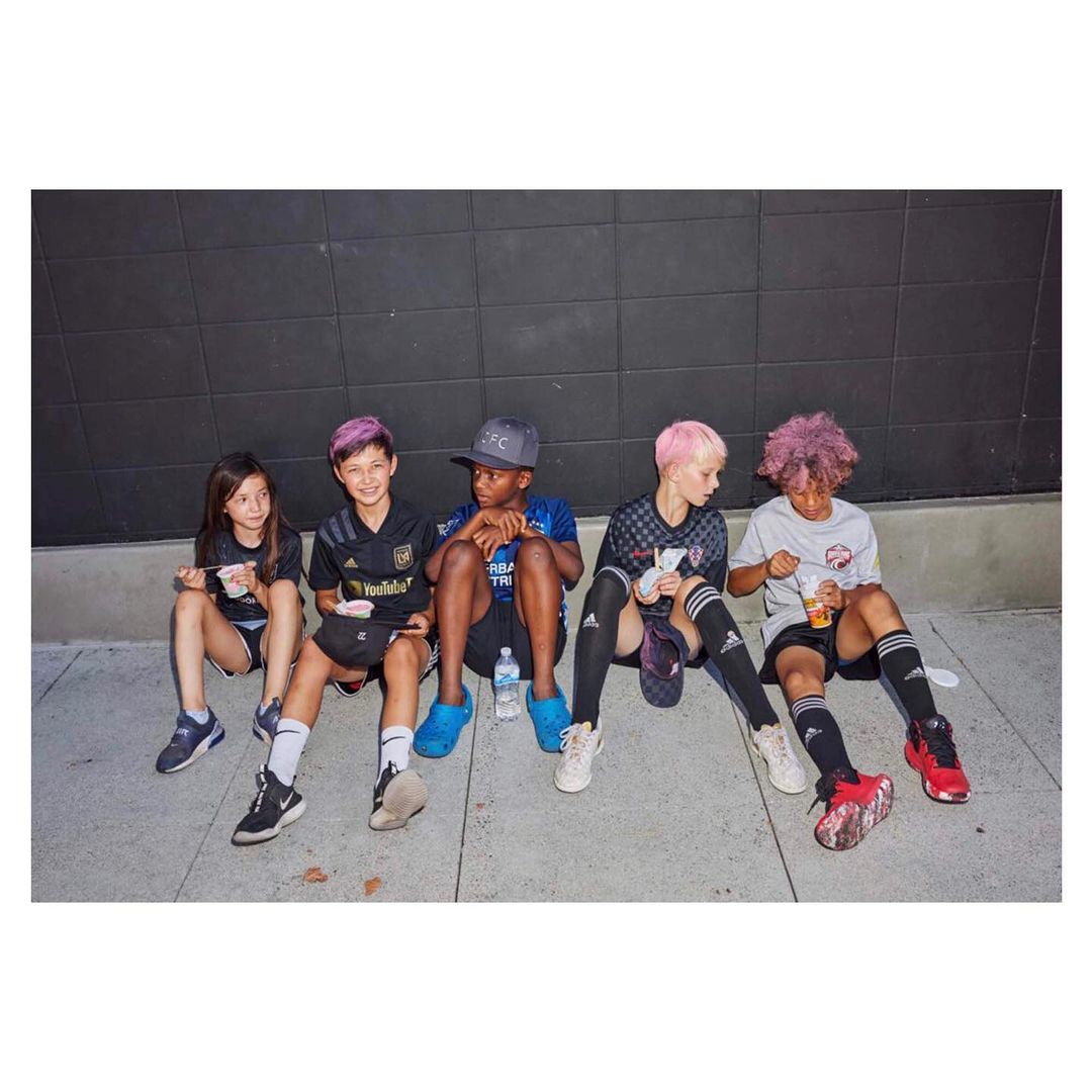 Group of five children in athletic wear sit against a dark wall; they appear to be resting after a game and are eating snacks.