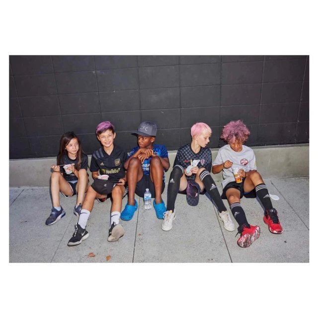 Group of five children in athletic wear sit against a dark wall; they appear to be resting after a game and are eating snacks.