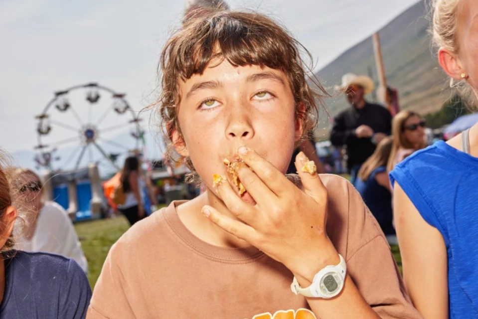 A girl with brown hair and eyes rolled back is eating food off her fingers at an outdoor fair with a Ferris wheel and people in background.