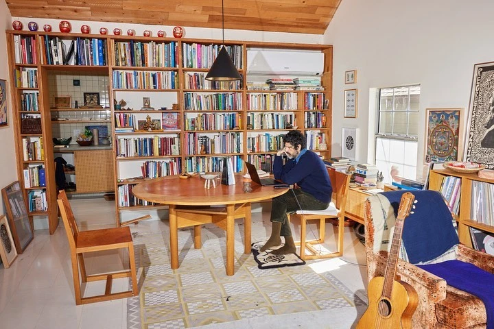Man works from home at a wooden table in front of a large bookshelf; a guitar leans against a chair in the foreground.