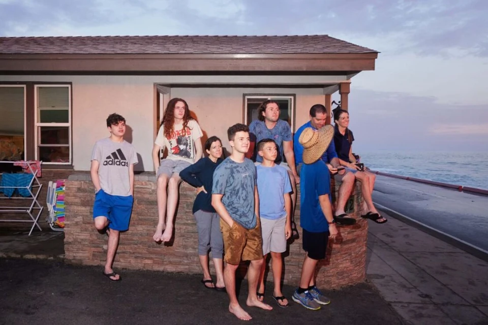 Group portrait of eight people outside a beach house at dusk. A mix of adults and children pose near the ocean with a dramatic sky.