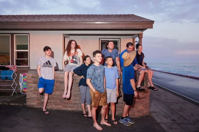 Group portrait of eight people outside a beach house at dusk. A mix of adults and children pose near the ocean with a dramatic sky.