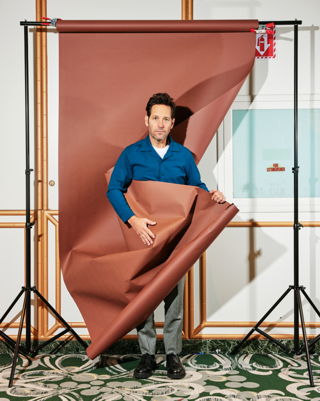 Paul Rudd holding backdrop paper in a photo studio, wearing a blue collared shirt and grey pants, standing on a patterned carpet.