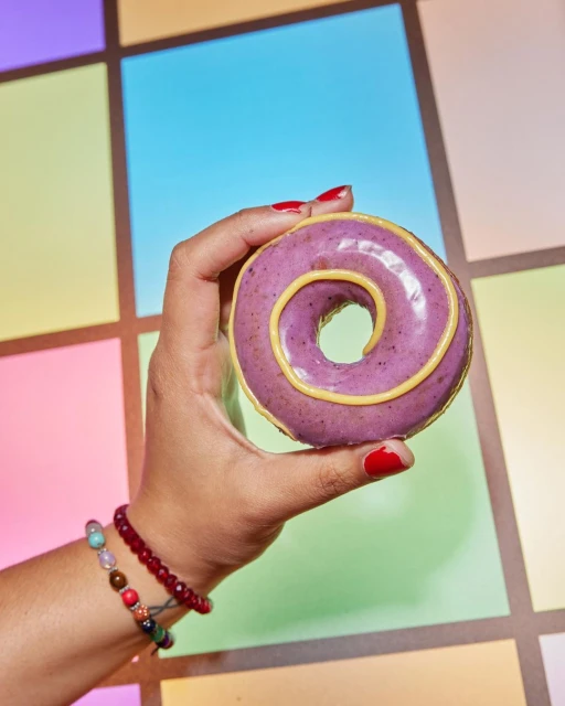 Hand holding a purple doughnut with yellow icing in front of a colorful tiled background in an eye-level, medium studio shot.