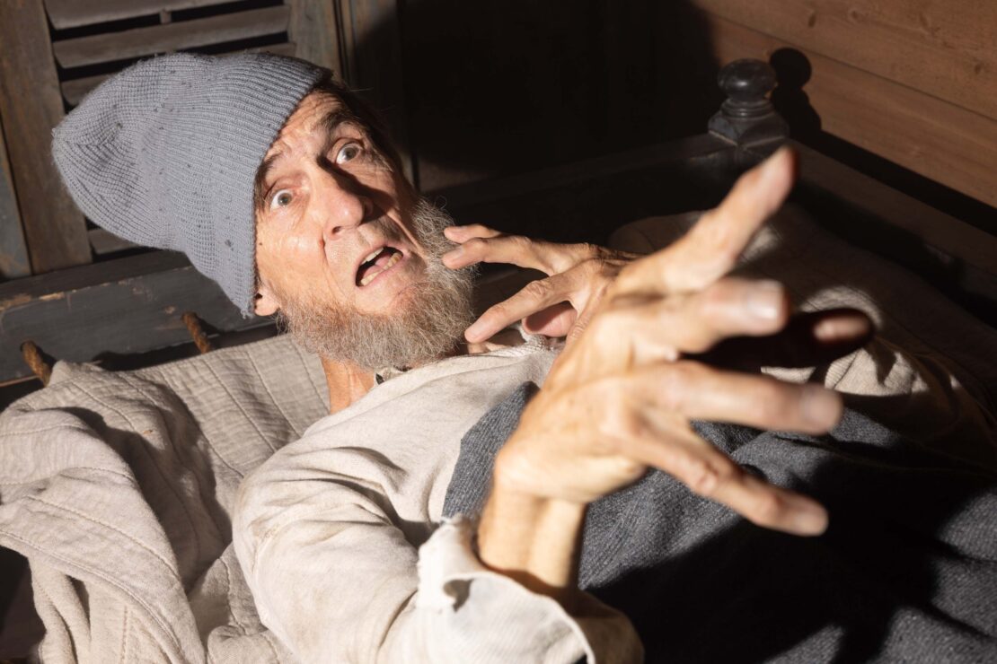 Elderly man with a gray beard and cap lies in bed with a scared facial expression, reaching out with his hands in a dimly lit room.