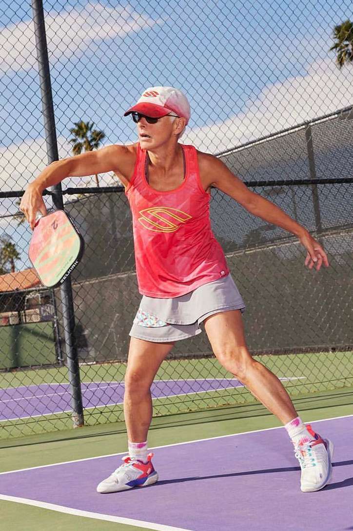 A fair-skinned woman in sportswear plays pickleball on a purple court, holding a paddle, and wearing a visor and sunglasses outdoors.